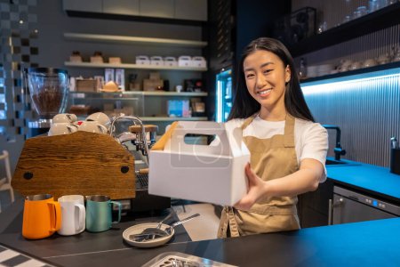 Cheerful coffee shop worker holding in her hands takeaway food packed in the cardboard box