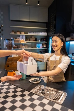 Photo for Cheerful female employee handing a takeout dessert packed in the cardboard box to a cafe client - Royalty Free Image