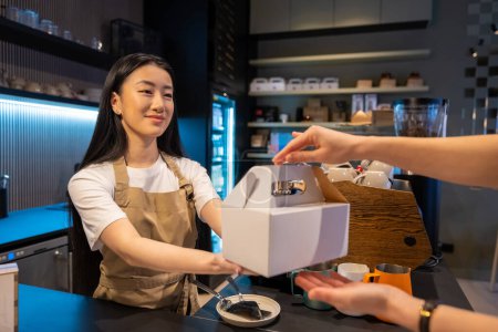 Téléchargez les photos : Souriant jeune ouvrier de café distribuant des plats à emporter emballés dans la boîte en carton à un client - en image libre de droit