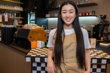 Foto de Sonriente lindo de pelo largo joven asiática empleada apoyada en el mostrador en la cafetería - Imagen libre de derechos