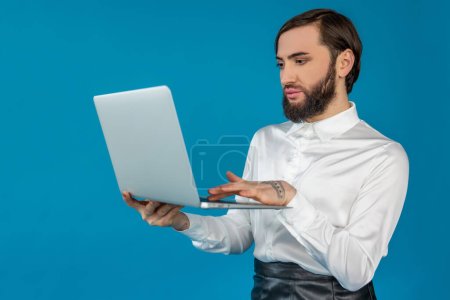Téléchargez les photos : Brunette homme transgenre gracieux portant une chemise blanche travaillant sur un ordinateur portable, tapant sur le clavier posant isolé sur fond bleu - en image libre de droit