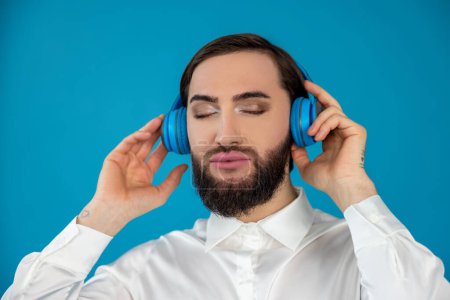 Téléchargez les photos : Satisfait homme transsexuel détendu en chemise blanche écoutant de la musique sur écouteurs profiter du temps libre avec les yeux fermés isolé sur fond bleu - en image libre de droit