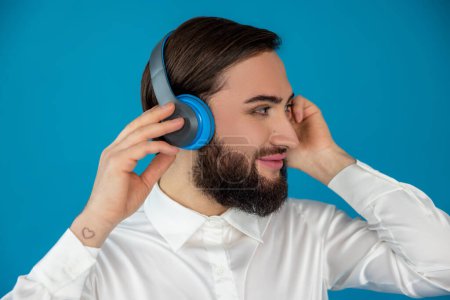 Foto de Perfil retrato de sonriente hombre barbudo transexual encantado escuchando música en auriculares mirando hacia otro lado aislado sobre fondo azul - Imagen libre de derechos
