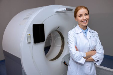 Jeune médecin blonde adulte debout près d'un scanner de tomodensitométrie dans un hôpital, vêtu d'un blouse de laboratoire, souriant à la caméra.