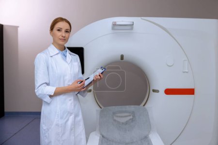 Téléchargez les photos : Médecin professionnel féminin en imagerie par résonance magnétique ou salle de tomodensitométrie d'un hôpital moderne, prenant des notes de diagnostic. - en image libre de droit