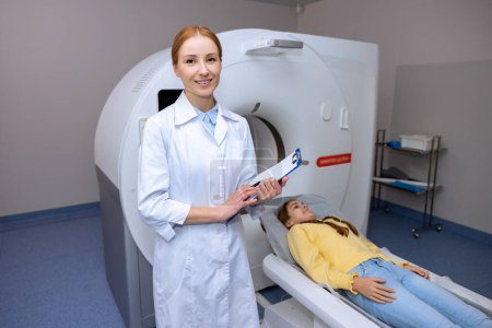 Photo for Little girl lying on table of mri scan machine while female radiographer starting modern health diagnostics. - Royalty Free Image