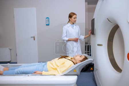 Foto de Niña acostada en la mesa de la máquina de escaneo de resonancia magnética mientras la radiografía femenina presiona el botón en el panel de control, diagnósticos de salud modernos. - Imagen libre de derechos