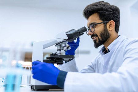Foto de Médico sonriente que trabaja en el científico moderno de investigación de laboratorio examina la sustancia bajo el microscopio, ajusta el espécimen en la placa de Petri. - Imagen libre de derechos
