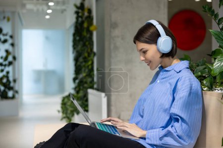 Photo for Smiling pleased cute young Caucasian woman in the wireless headphones typing on the portable computer - Royalty Free Image