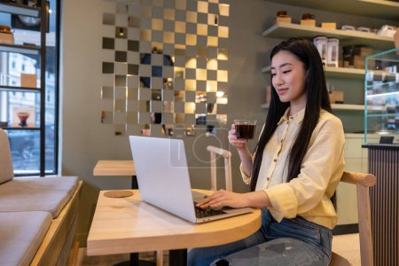 Téléchargez les photos : Agréable employée à distance avec une tasse de boisson caféinée assise à la table du café travaillant sur l'ordinateur portable - en image libre de droit