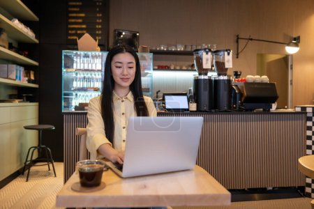 Photo for Concentrated young Asian remote worker sitting at the cafe table while typing on the laptop - Royalty Free Image
