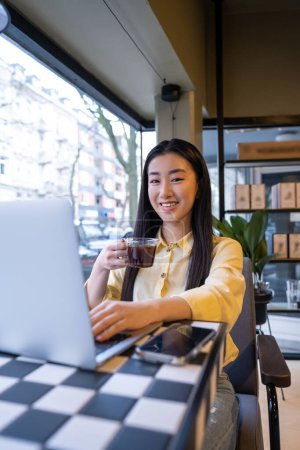 Foto de Alegre empleado remoto con una taza de vidrio de bebida con cafeína trabajando en la computadora portátil en la mesa de la cafetería - Imagen libre de derechos