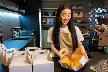 Photo for Waist-up portrait of a contented worker packing a slice of cream cake into the cardboard box at the counter - Royalty Free Image