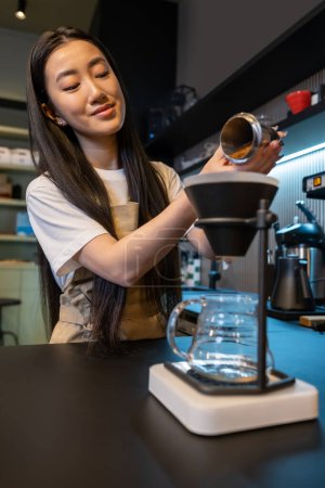 Photo for Focused Asian cafe worker pouring the ground coffee into the coffee maker funnel with filter - Royalty Free Image