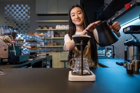 Photo for Waist-up portrait of a smiling barista tilting the kettle over the pour-over coffee maker funnel with filter - Royalty Free Image