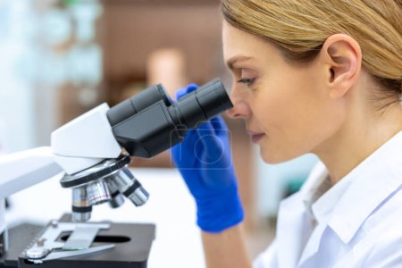 Foto de Laboratorio profesional. Retrato del perfil de una trabajadora médica rubia con una bata blanca y guantes, usando microscopio durante la investigación. - Imagen libre de derechos