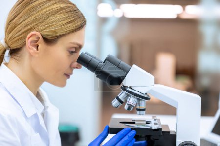 Téléchargez les photos : Portrait latéral d'une femme scientifique blonde regardant une cellule biochimique en laboratoire à l'aide d'un microscope, étudiant la biologie moléculaire. - en image libre de droit
