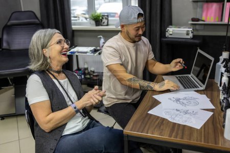 Foto de En el salón de tatuajes. Mujer de pelo gris sentada con un maestro del tatuaje - Imagen libre de derechos