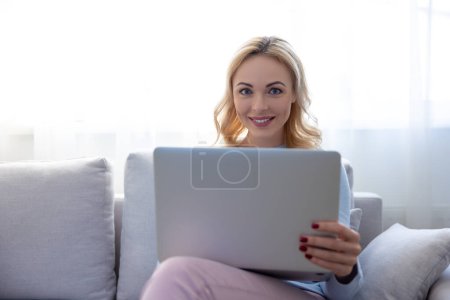 Photo for Pleased pretty blonde lady seated on the cozy sofa with her laptop looking before her - Royalty Free Image