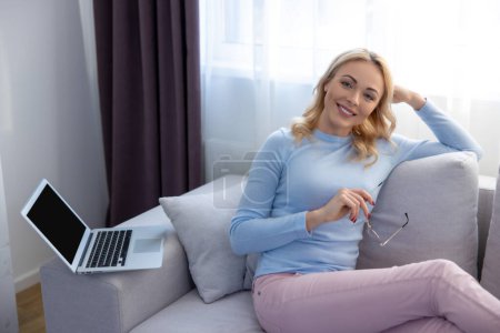 Photo for Smiling happy lady sitting on the sofa with a pair of eyeglasses in the hand - Royalty Free Image