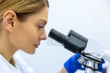 Foto de Mujer rubia asistente de laboratorio en guantes azules mira en un microscopio, fórmula química en sala de pruebas de laboratorio, mirando célula bioquímica en laboratorio. - Imagen libre de derechos