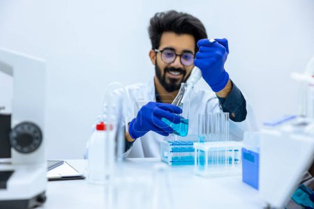 Téléchargez les photos : Jeune chimiste barbu souriant dans des gants de protection assis à table et tenant une fiole d'échantillon médical, travaillant avec des liquides. - en image libre de droit
