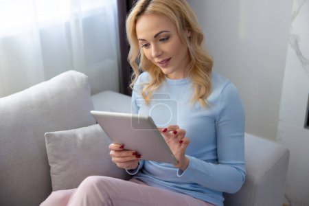 Téléchargez les photos : Focalisé blonde jeune femme assise sur le canapé confortable tout en tapant sur sa tablette numérique - en image libre de droit