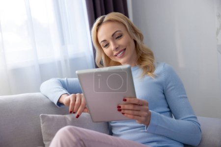 Téléchargez les photos : Femme souriante assise sur le canapé confortable regardant la tablette numérique dans ses mains - en image libre de droit