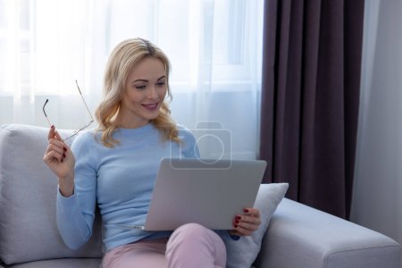 Foto de Mujer rubia concentrada sentada en el sofá mirando una tableta en sus manos - Imagen libre de derechos