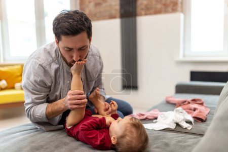 Photo for Young caucasian father kissing feet of his infant baby daughter at home interior. - Royalty Free Image