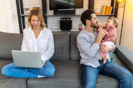Photo for Father carrying infant daughter while mother using laptop on sofa remote work husband helping his wife with baby child. - Royalty Free Image