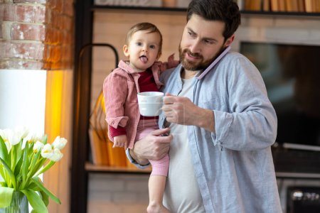 Foto de Feliz padre multitarea con pequeño niño llamando en el teléfono inteligente en casa y beber café. - Imagen libre de derechos