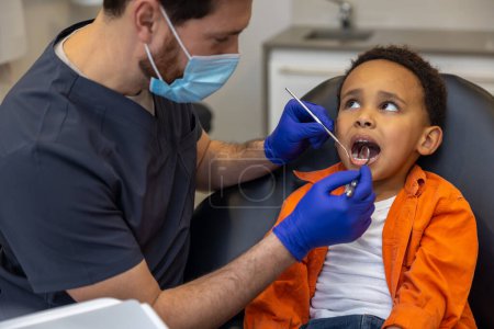 Foto de En odontología. Niño de piel oscura mirando asustado mientras el médico revisa sus dientes - Imagen libre de derechos