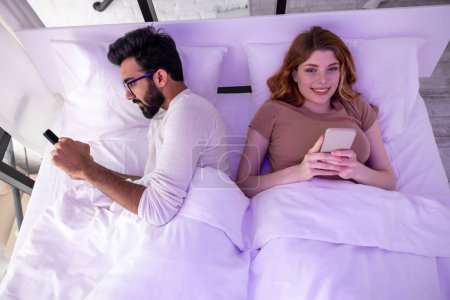 Foto de Pareja joven sonriendo esposa y marido en la cama usando teléfonos ignorándose mutuamente. - Imagen libre de derechos