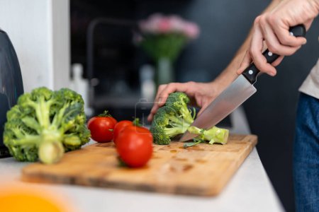 Photo for Unknown male hand cuts tomato and brocoli on cutting board with sharp knife in kitchen, making salad - Royalty Free Image
