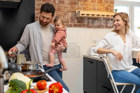 Photo for Happy family at home in the kitchenn man with baby cooking making meal woman sitting at table. - Royalty Free Image