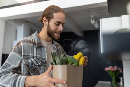 Photo for Satisfied man ordered food delivery to his home, unpacking online order of vegetables and fruits from store, fast delivery of fresh products. - Royalty Free Image
