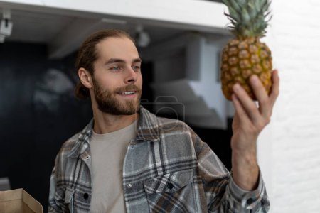Photo for Joyful bearded man in checkered shirt holding pineapples in hands standing in kitchen at home. - Royalty Free Image