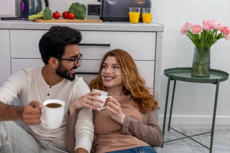 Foto de Esposa y marido sentados en el suelo en la cocina y hablando, bebiendo café de la mañana en casa. - Imagen libre de derechos