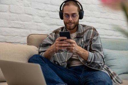 Photo for Smiling young man wearing headphones listen to mobile music playing in smartphone app, relaxing holding using phone sitting on sofa at home. - Royalty Free Image