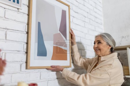 Téléchargez les photos : Design d'intérieur. Femme âgée mettre une photo sur le mur à la maison - en image libre de droit