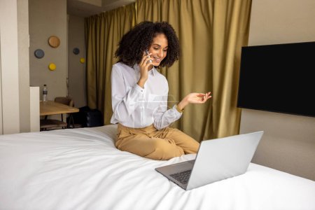 Foto de En el hotel. Mujer de negocios de pelo rizado hablando por teléfono en la habitación de hotel - Imagen libre de derechos