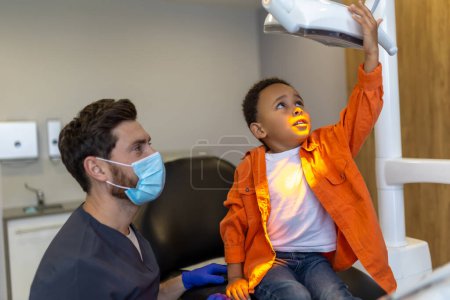 Téléchargez les photos : En dentisterie. Garçon à la peau foncée loking intéressé par la lampe dentaire - en image libre de droit
