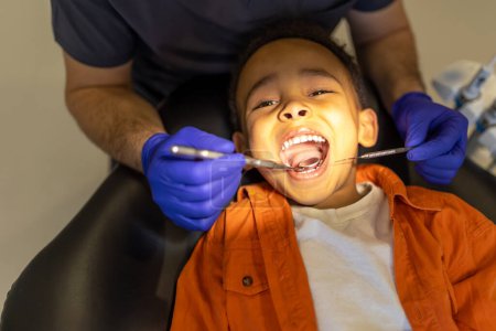 Téléchargez les photos : Chez les dentistes. Dentiste examinant la cavité buccale d'un petit garçon à la peau foncée - en image libre de droit