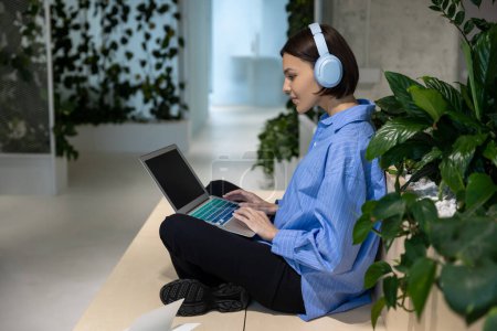 Téléchargez les photos : Vue latérale d'une dame concentrée dans les écouteurs assis dans un espace de coworking en regardant l'écran de l'ordinateur portable - en image libre de droit
