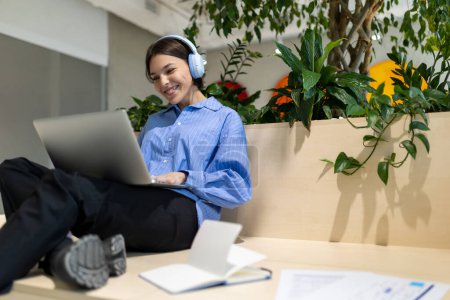 Photo for Cheerful young female in the wireless headphones seated in a coworking space looking at the computer monitor - Royalty Free Image