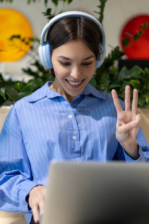 Photo for Joyous lady in the headphones showing a three-finger gesture while looking at the laptop screen - Royalty Free Image