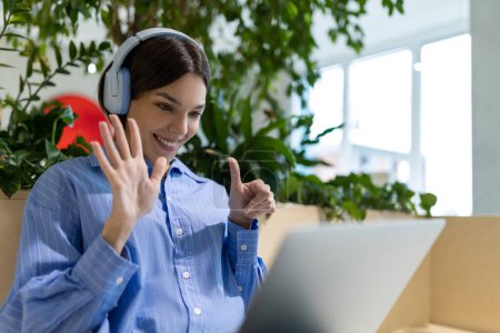 Photo for Waist-up portrait of a smiling young female in the headphones showing the thumbs-up gesture while looking at the laptop screen - Royalty Free Image