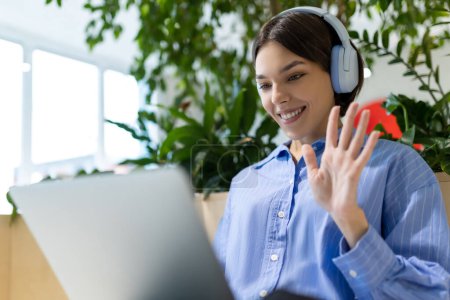 Photo for Waist-up portrait of a cheerful woman in the headphones waving at someone while looking at the computer monitor - Royalty Free Image