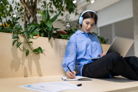 Foto de Mujer seria enfocada en los auriculares sentados en un espacio de coworking haciendo notas en el cuaderno - Imagen libre de derechos